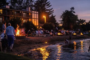 A beachside gathering with people around fires, a building in the background, and a calm water body reflecting lights. - Cragun's Resort & Hotel (Brainerd)