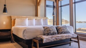 A bedroom with a large bed, a bench, and a view of a body of water through large windows. - Cragun's Resort & Hotel (Brainerd)