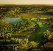 An aerial view of a golf course with a pond, surrounded by dense forest, and a winding road.