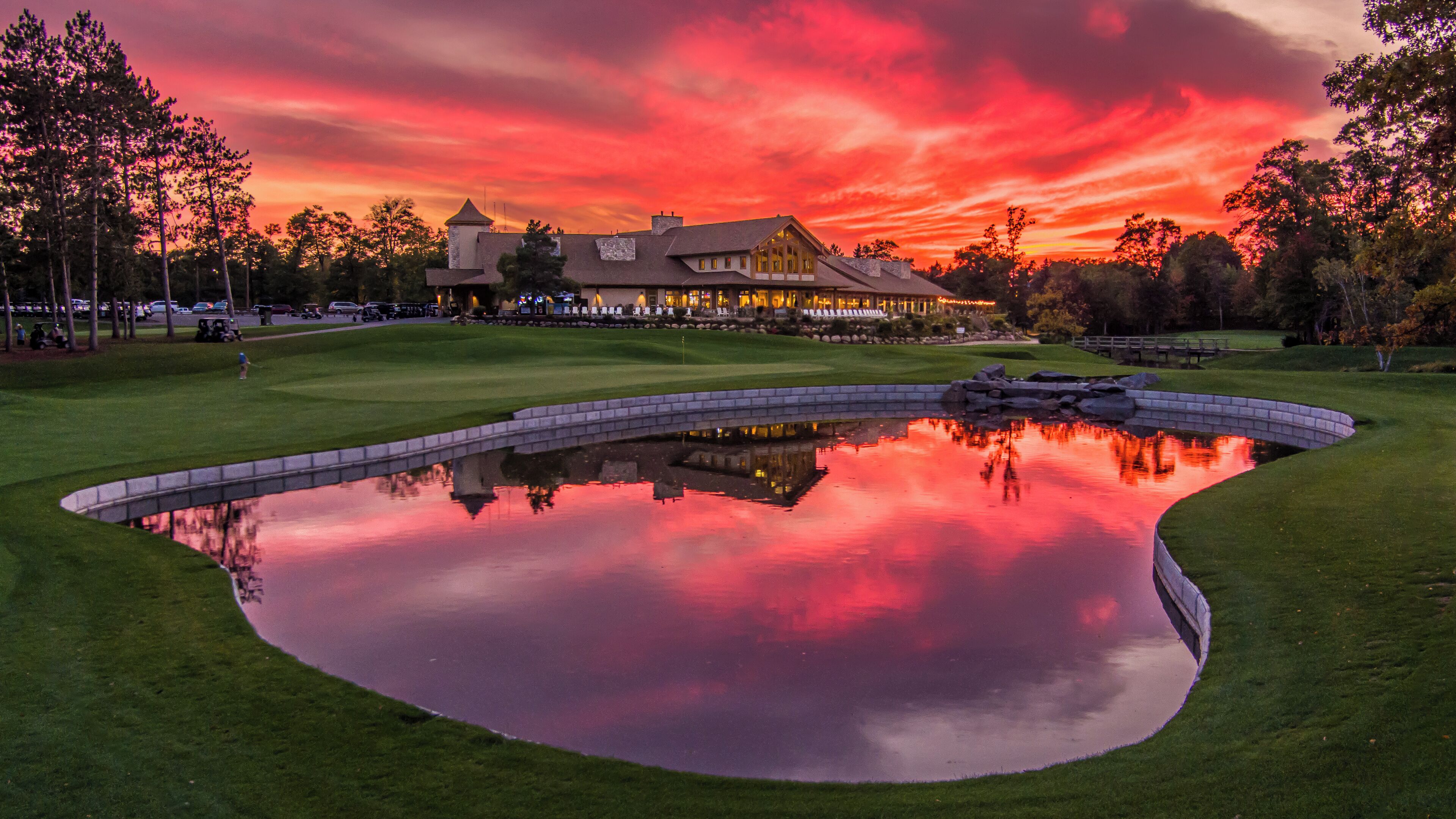 Un parcours de golf avec un étang reflétant le ciel au coucher du soleil, un club-house et un pont.