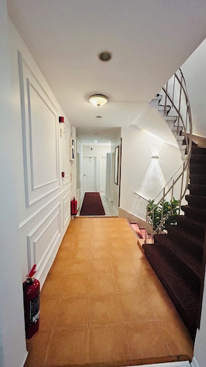 A hallway with a brown tiled floor, white walls, a red fire extinguisher, and a spiral staircase with a white railing. - Anadolu Hotel (Istanbul)
