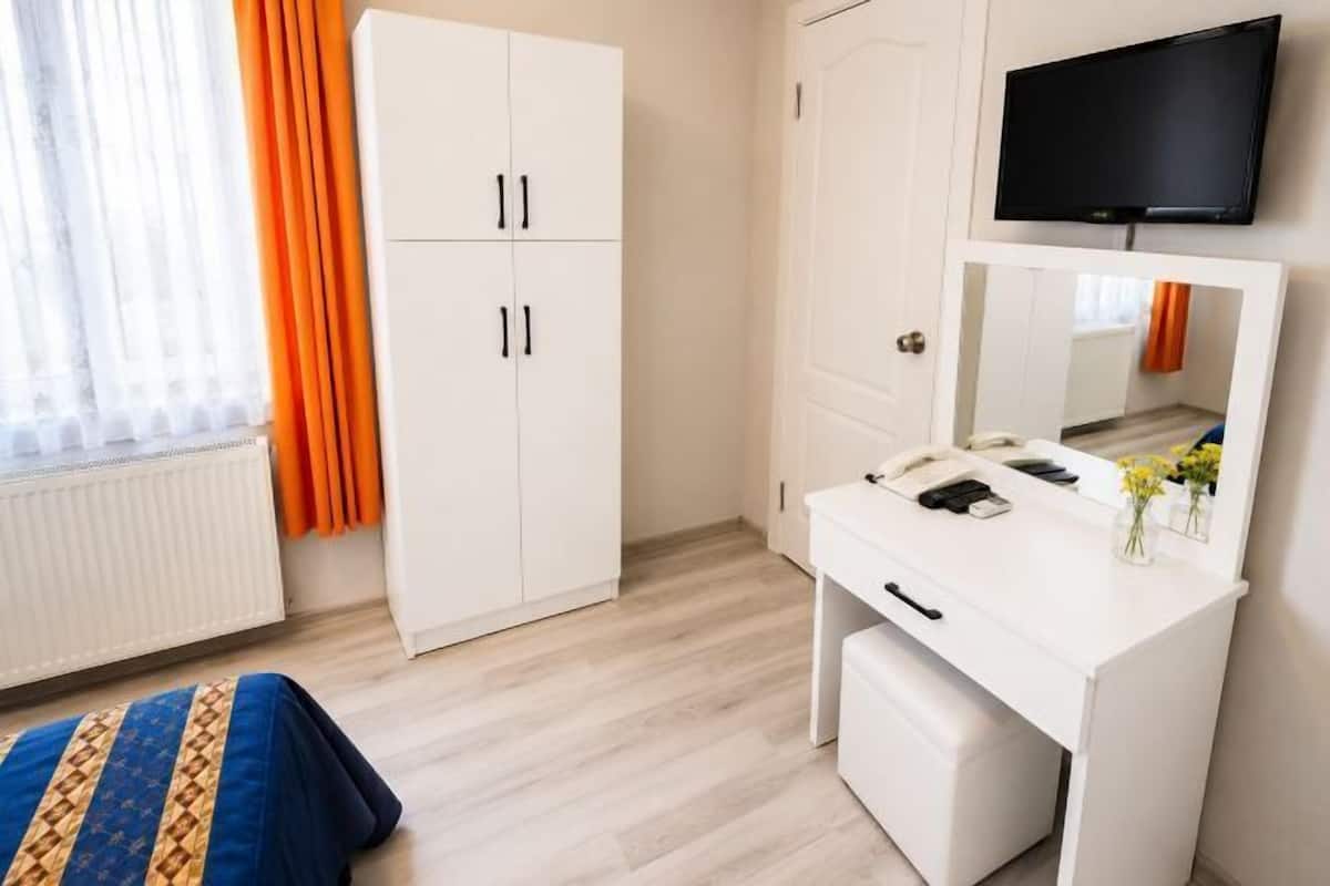 A room with a white wardrobe, a white desk with a mirror, a television mounted on the wall, and a window with orange curtains.