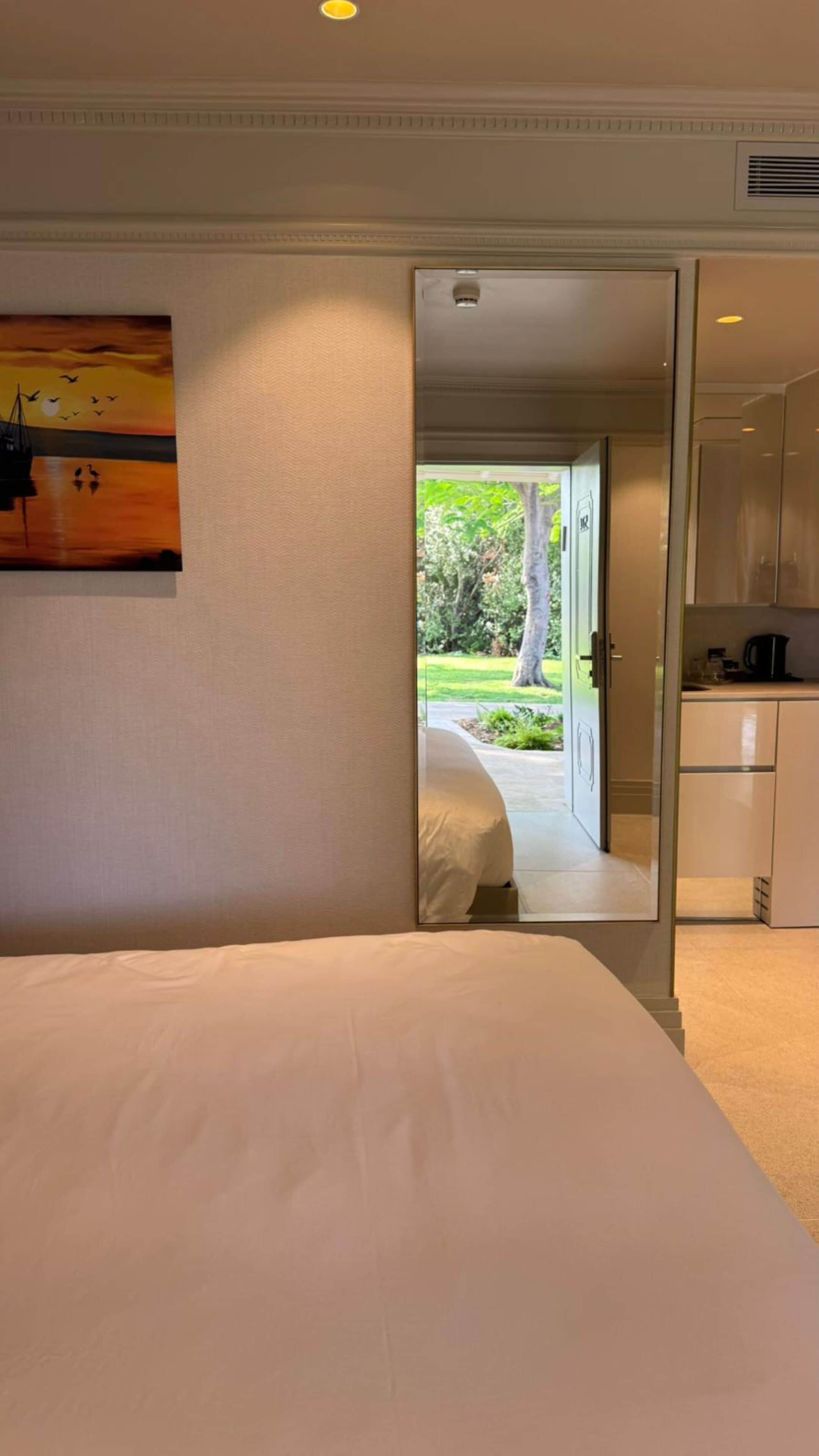 A hotel room with a bed, a painting, a view through an open door to a garden, and a kitchenette.