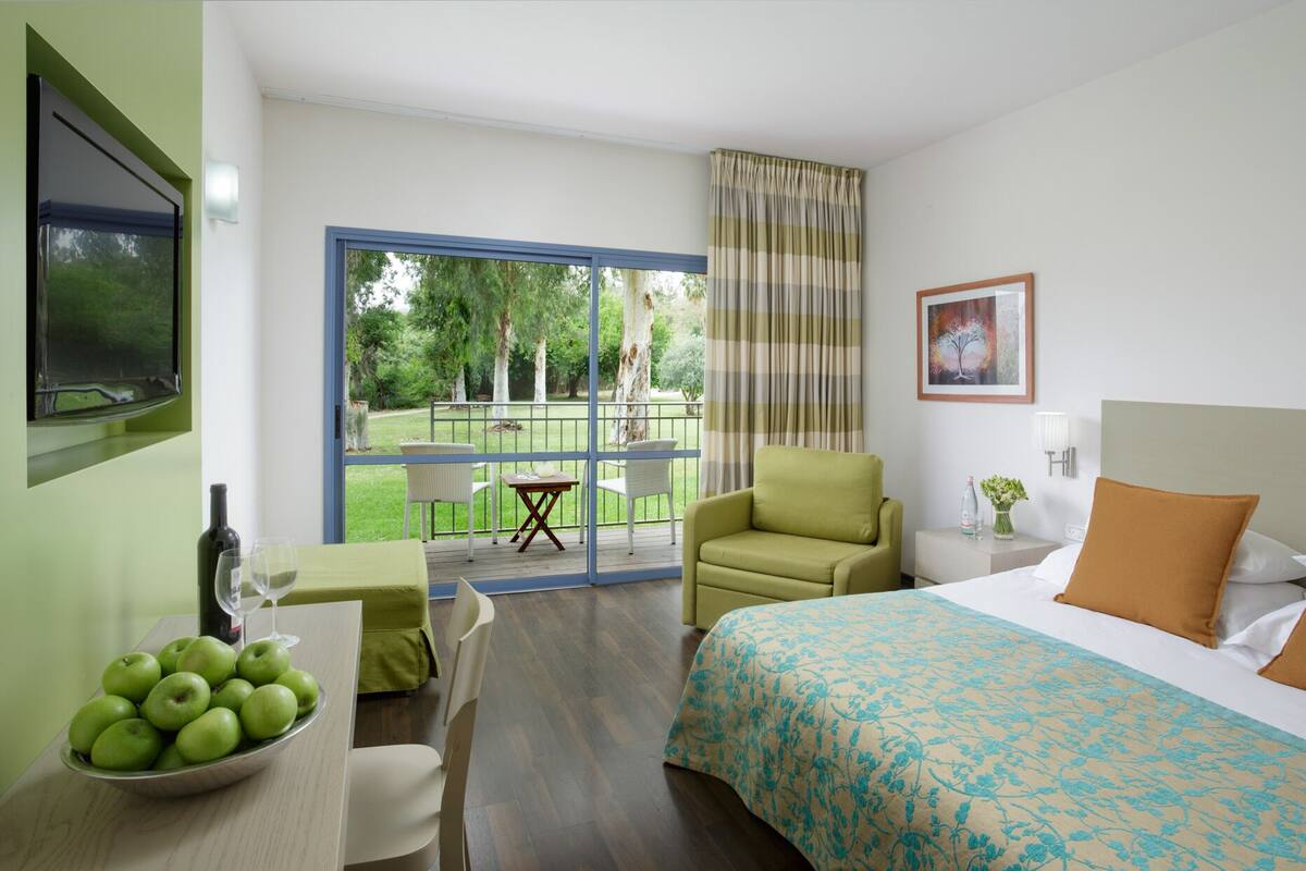 Ein Hotelzimmer mit Bett, Schreibtisch mit Apfelschale, Stuhl, kleinem Tisch und Blick auf einen Garten durch eine Glastür.