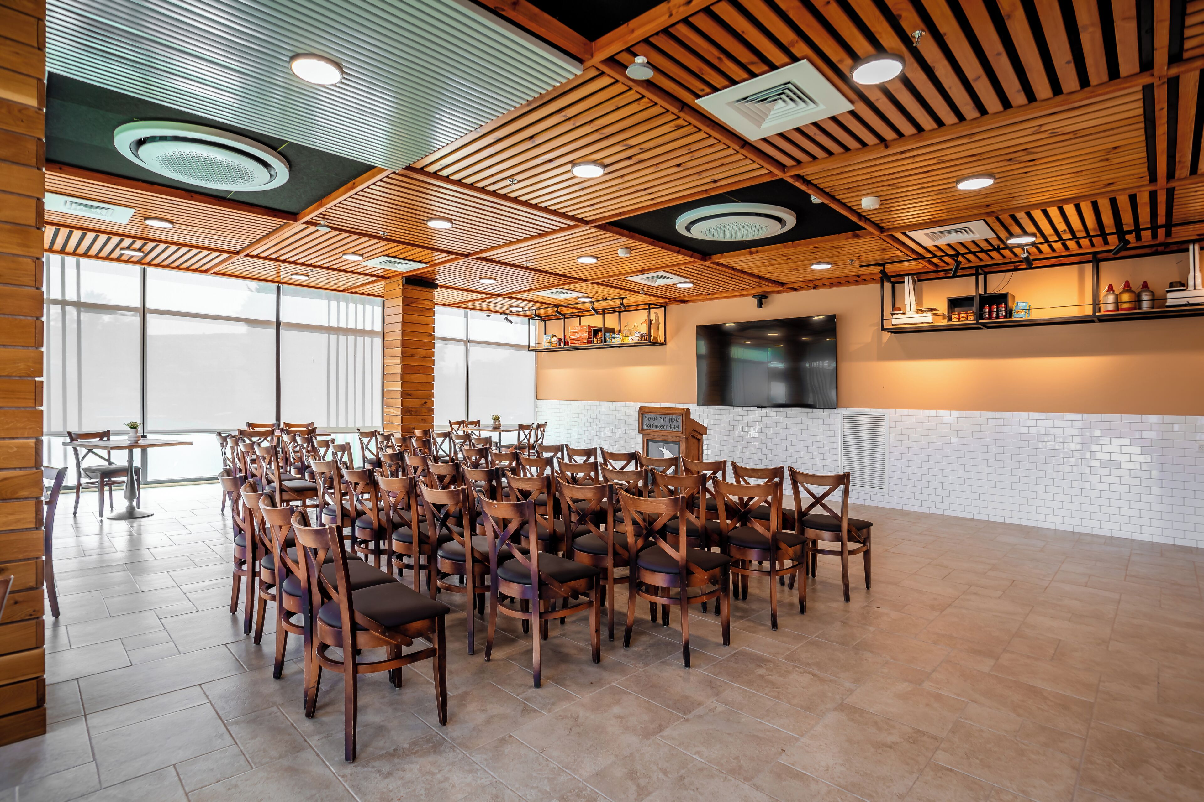 A spacious indoor area with wooden ceiling beams, a kitchen area with a counter and appliances, and multiple wooden chairs arranged for seating.