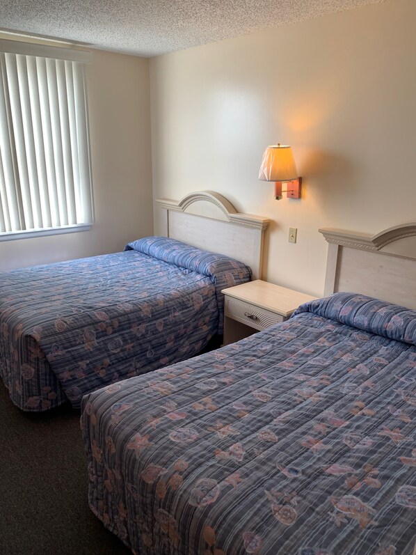 Habitación de hotel con dos camas, una mesita de noche, una lámpara y una ventana con persianas.