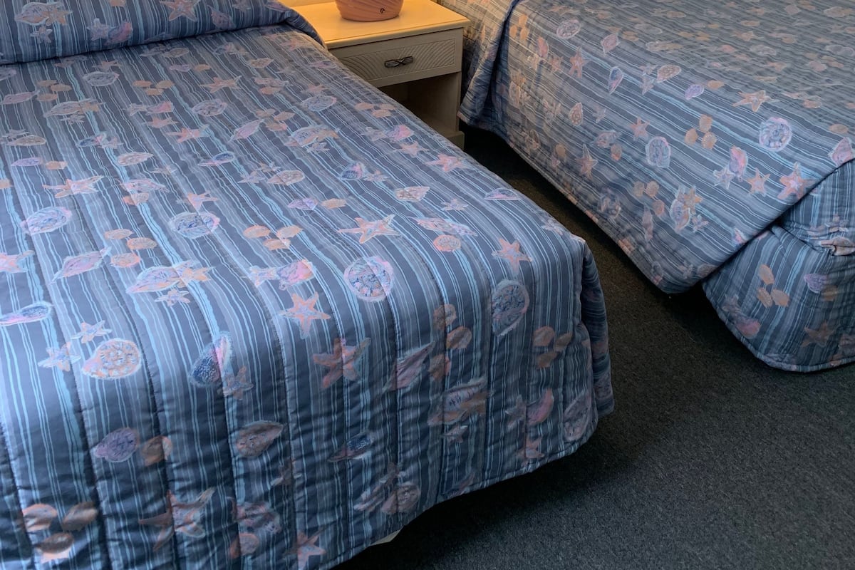 Hotellihuone, jossa on kaksi sänkyä. Jokaisessa sängyssä on sininen, kukkakuviollinen päiväpeite. Huoneessa on myös yöpöytä, jossa on lamppu, sekä seinään kiinnitetty valaisin.