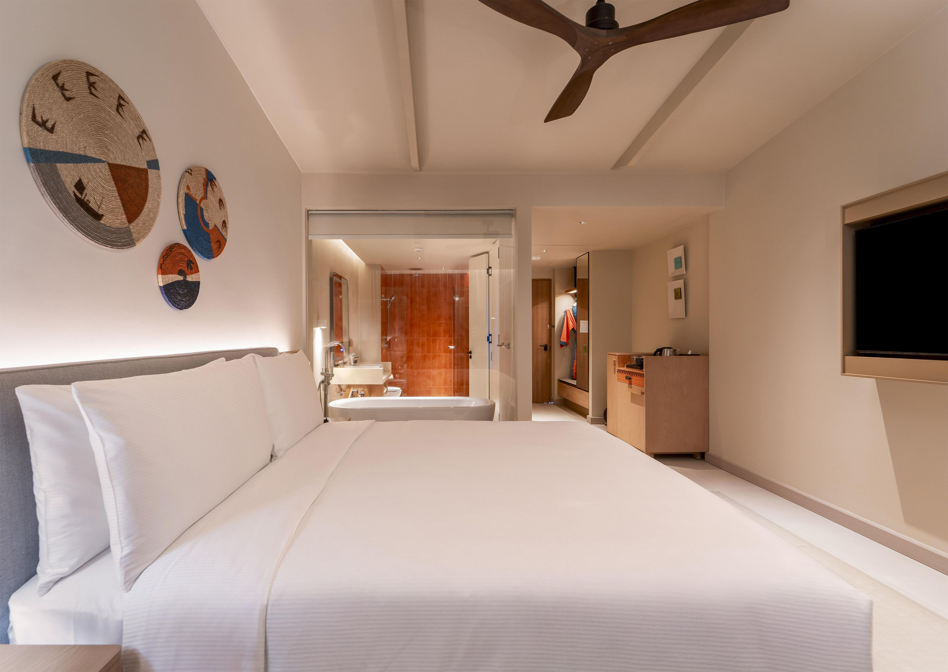 Una habitación de hotel con una cama grande, un ventilador de techo de madera, un baño con ducha y un televisor montado en la pared.