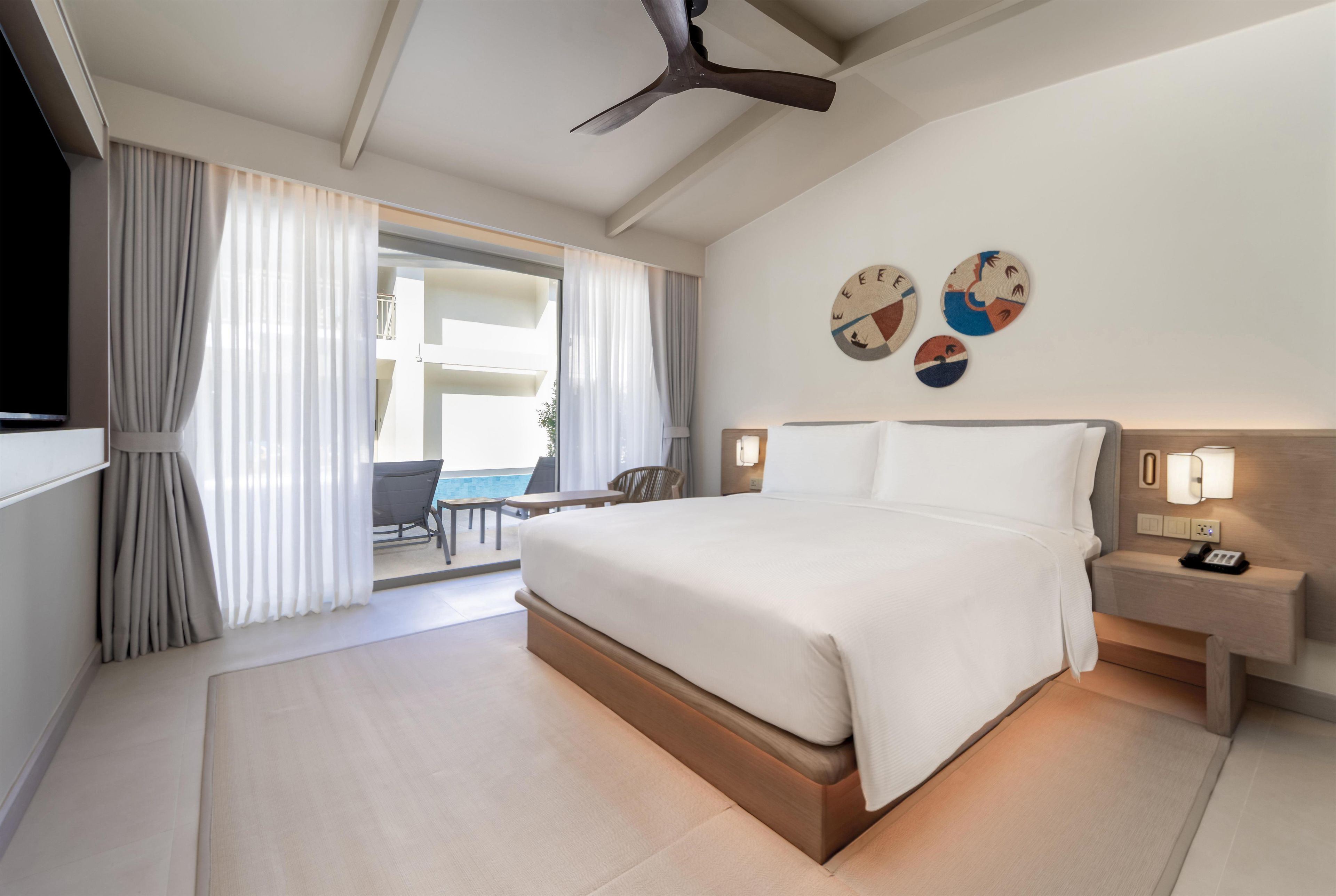 Habitación de hotel con una cama grande, ventilador de techo, escritorio con silla y balcón con cortinas.