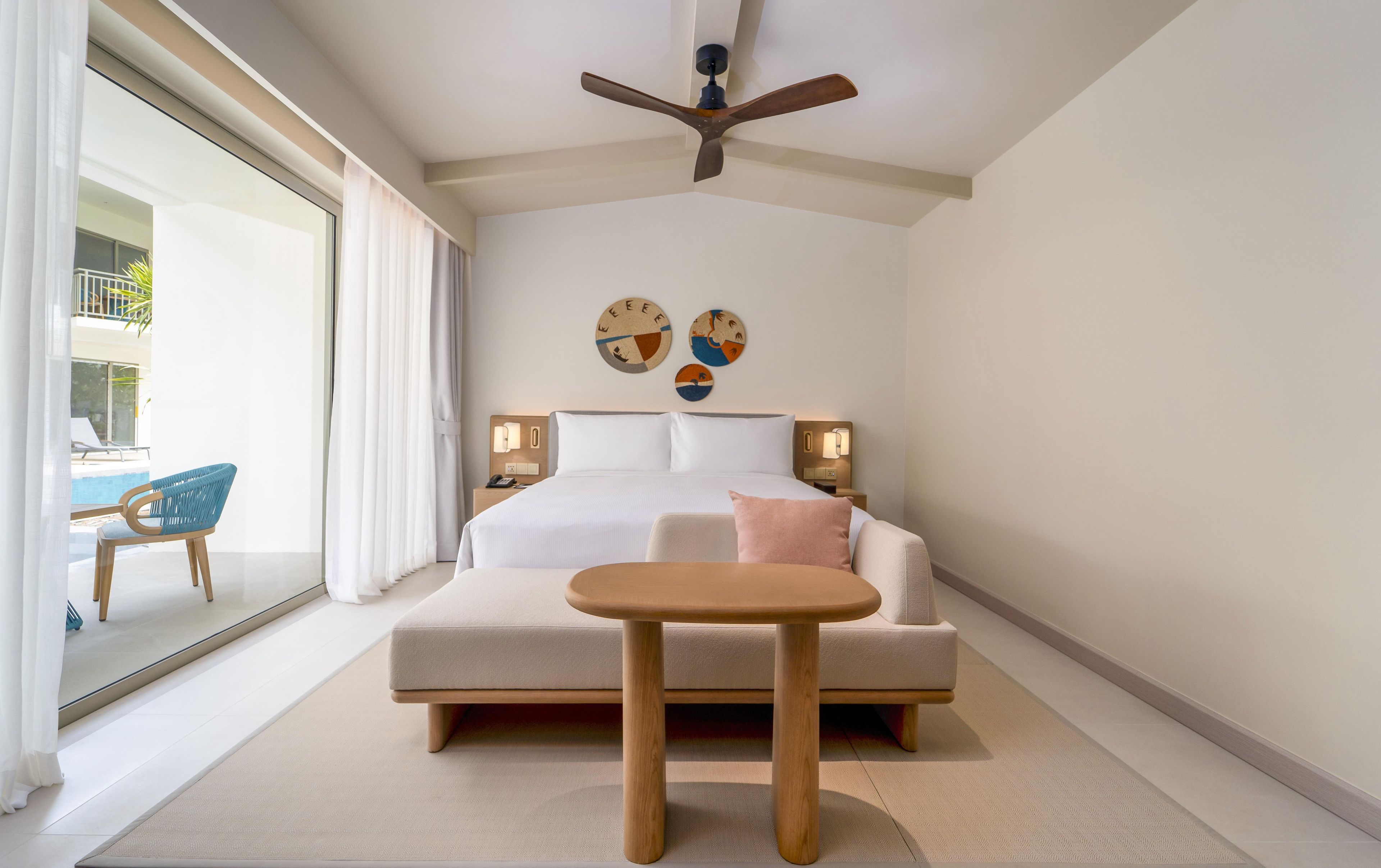 A hotel room with a bed, bedside tables, a ceiling fan, and a view of the pool area.