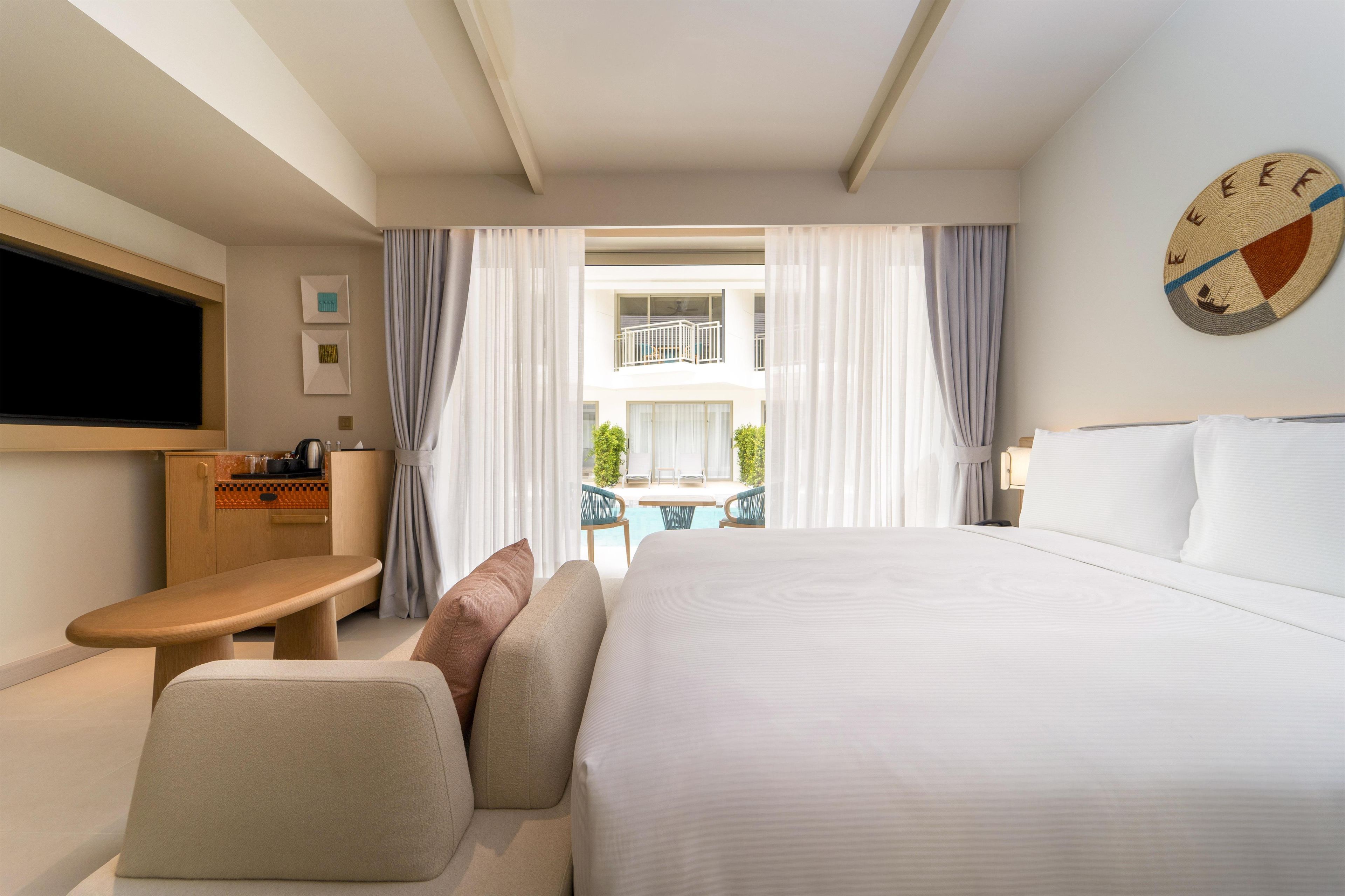 Habitación de hotel con cama, televisión, escritorio y vistas a un balcón con mobiliario exterior.