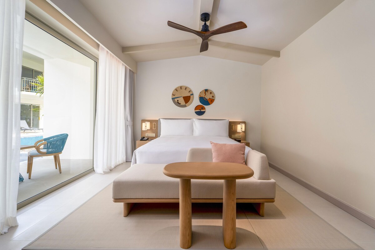 A hotel room with a bed, bedside tables, a ceiling fan, and a view of the pool area.