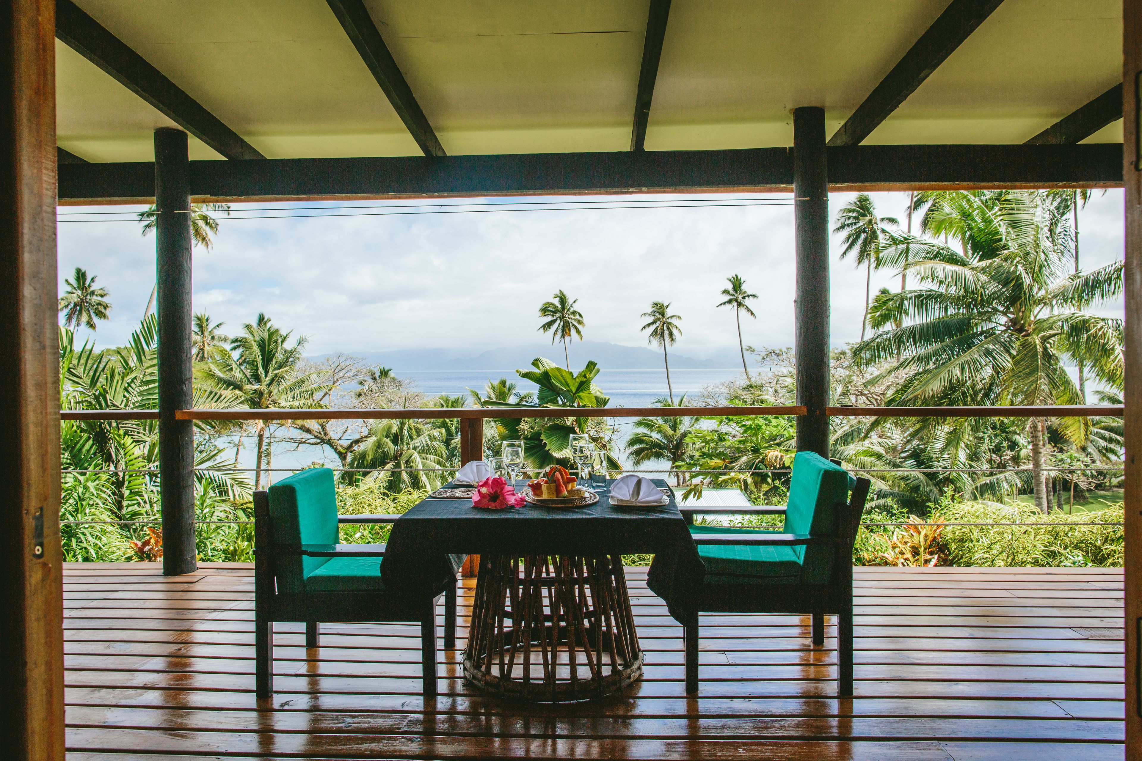 Un área de comedor al aire libre con techo, con una mesa lista para dos, con vistas a un paisaje tropical de palmeras y al océano.