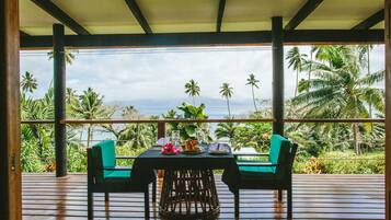 Un área de comedor al aire libre con techo, con una mesa lista para dos, con vistas a un paisaje tropical de palmeras y al océano.