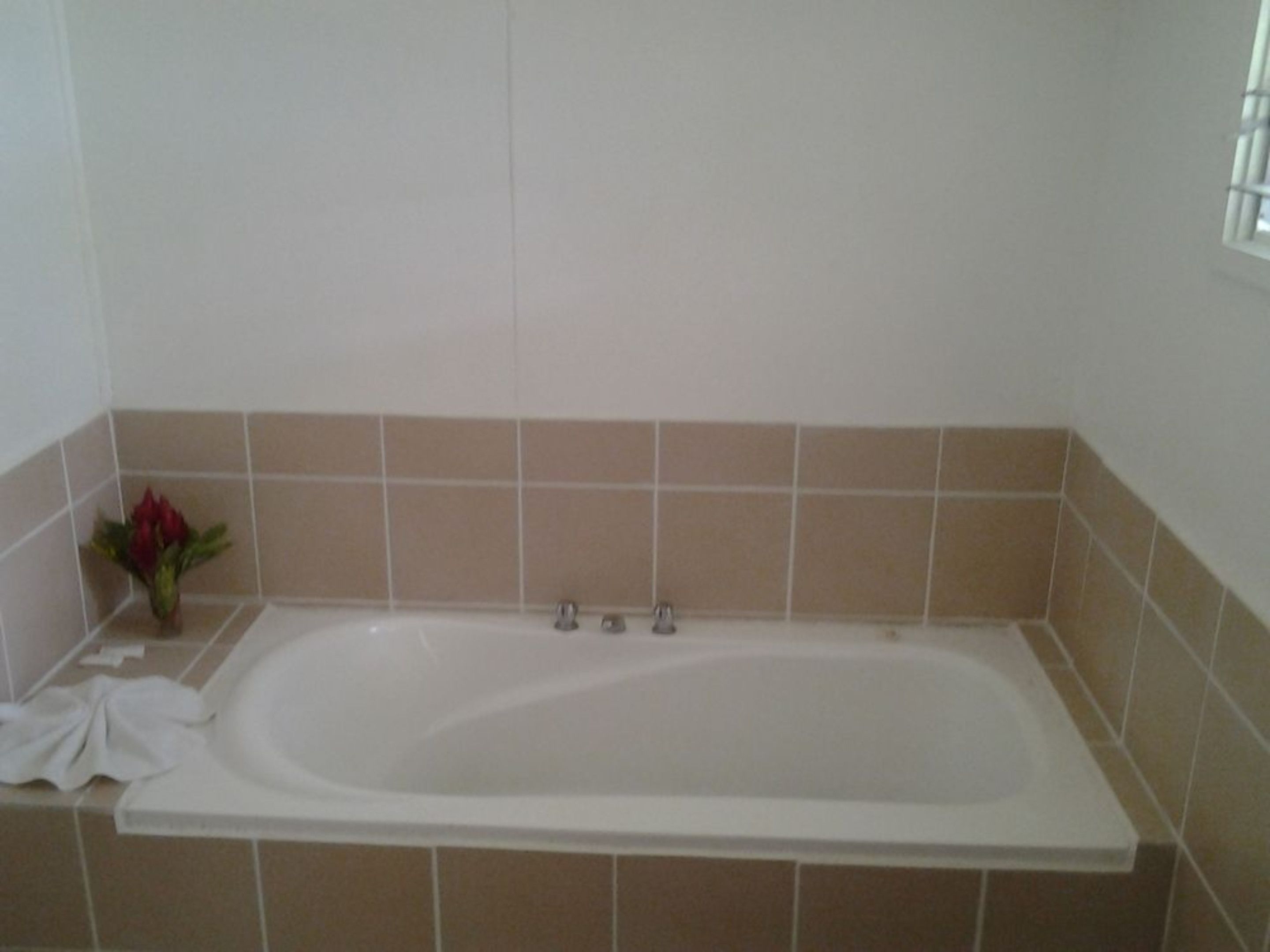 A standard rectangular bathtub with a white interior, beige tiled walls, and a small vase with a red flower on the left side.