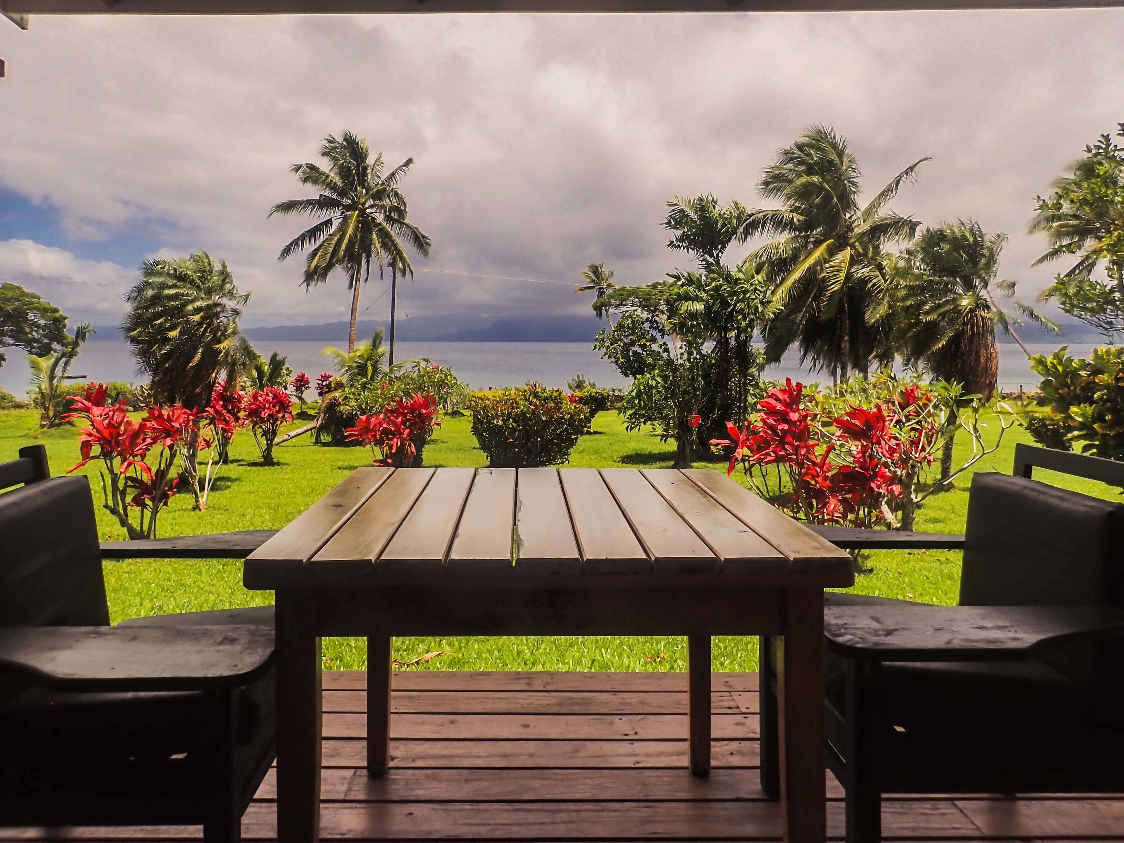 A wooden table with two chairs on a deck, overlooking a lush garden with palm trees and red flowers, and a view of the sea under a cloudy sky.
