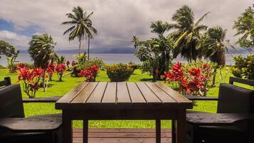 A wooden table with two chairs on a deck, overlooking a lush garden with palm trees and red flowers, and a view of the sea under a cloudy sky.