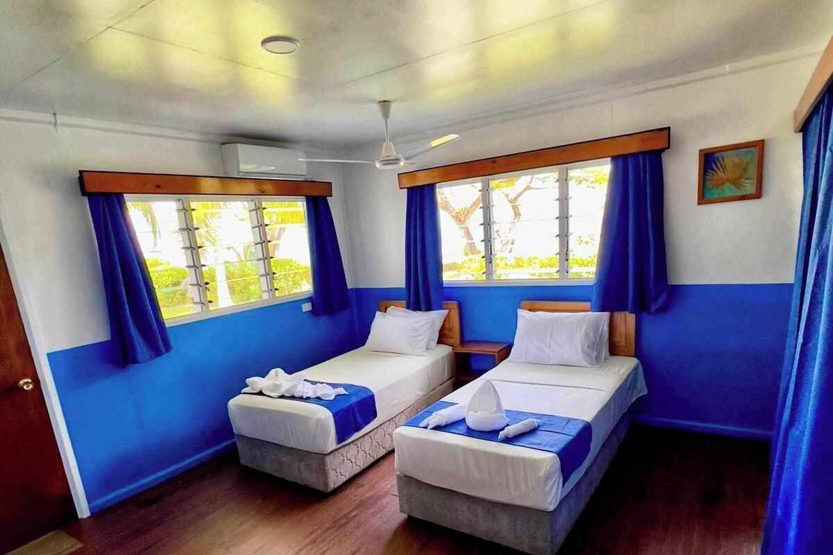 A room with two beds, wooden flooring, and large windows with blue curtains.