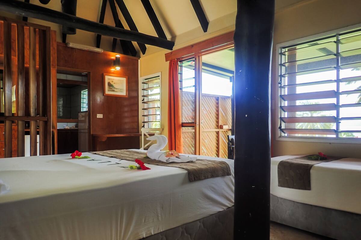 A hotel room with two beds, wooden paneling, a large window with blinds, and a view of greenery outside.