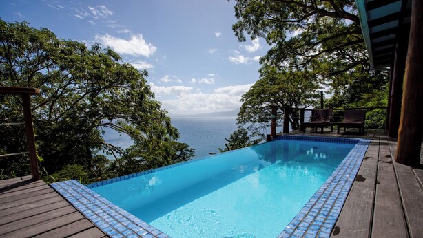 A clear blue infinity pool with a wooden deck and tiled edge, surrounded by lush greenery and overlooking a body of water. - Daku Resort (Savusavu)