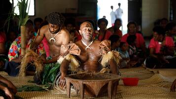 A group of people seated on woven mats, some wearing traditional attire with feathers and necklaces.