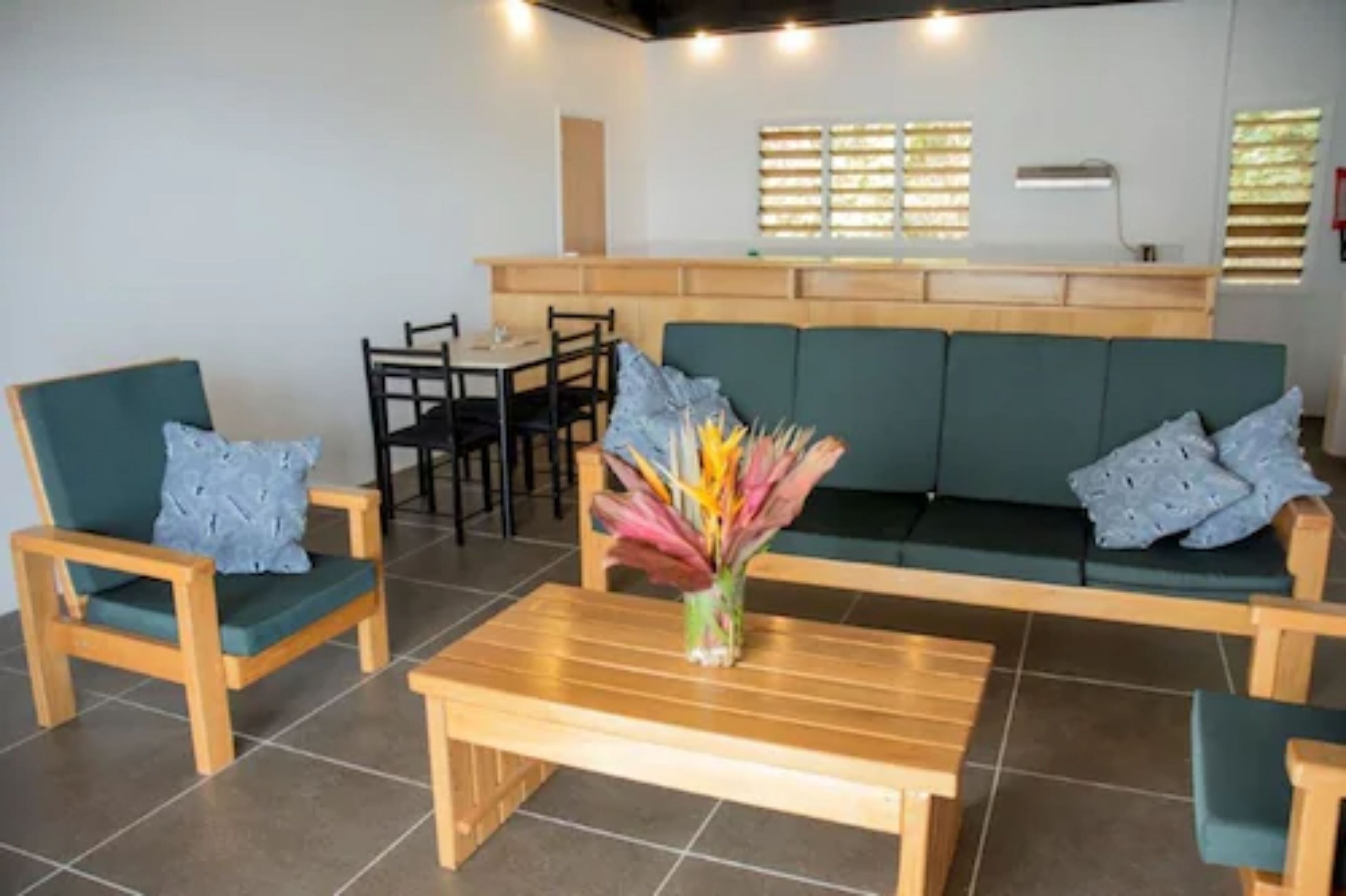 A room with a green sofa, two wooden chairs, a wooden table, and a vase with colorful flowers.
