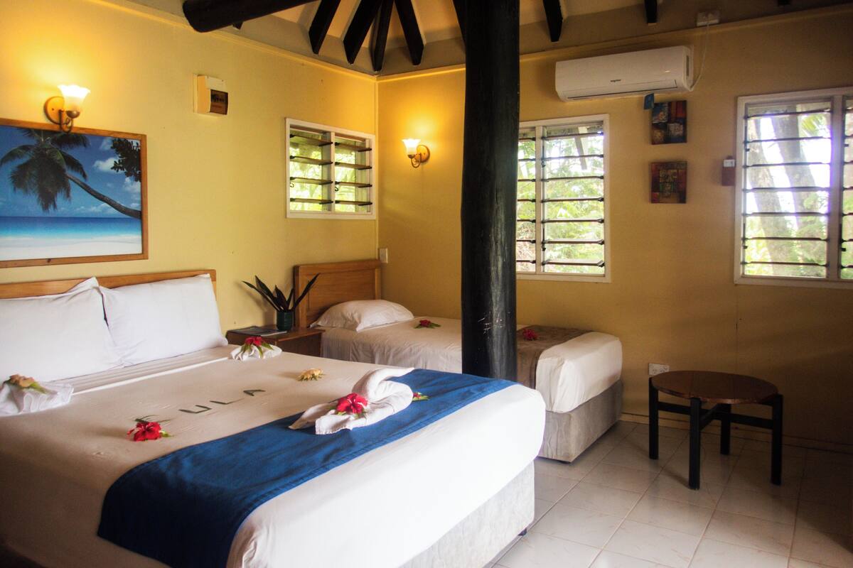 A hotel room with two beds, a wooden headboard, a potted plant, and a painting of a palm tree.