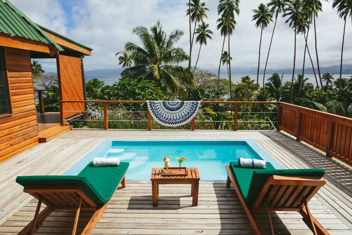 A wooden deck with a pool, lounge chairs, and a table with drinks.