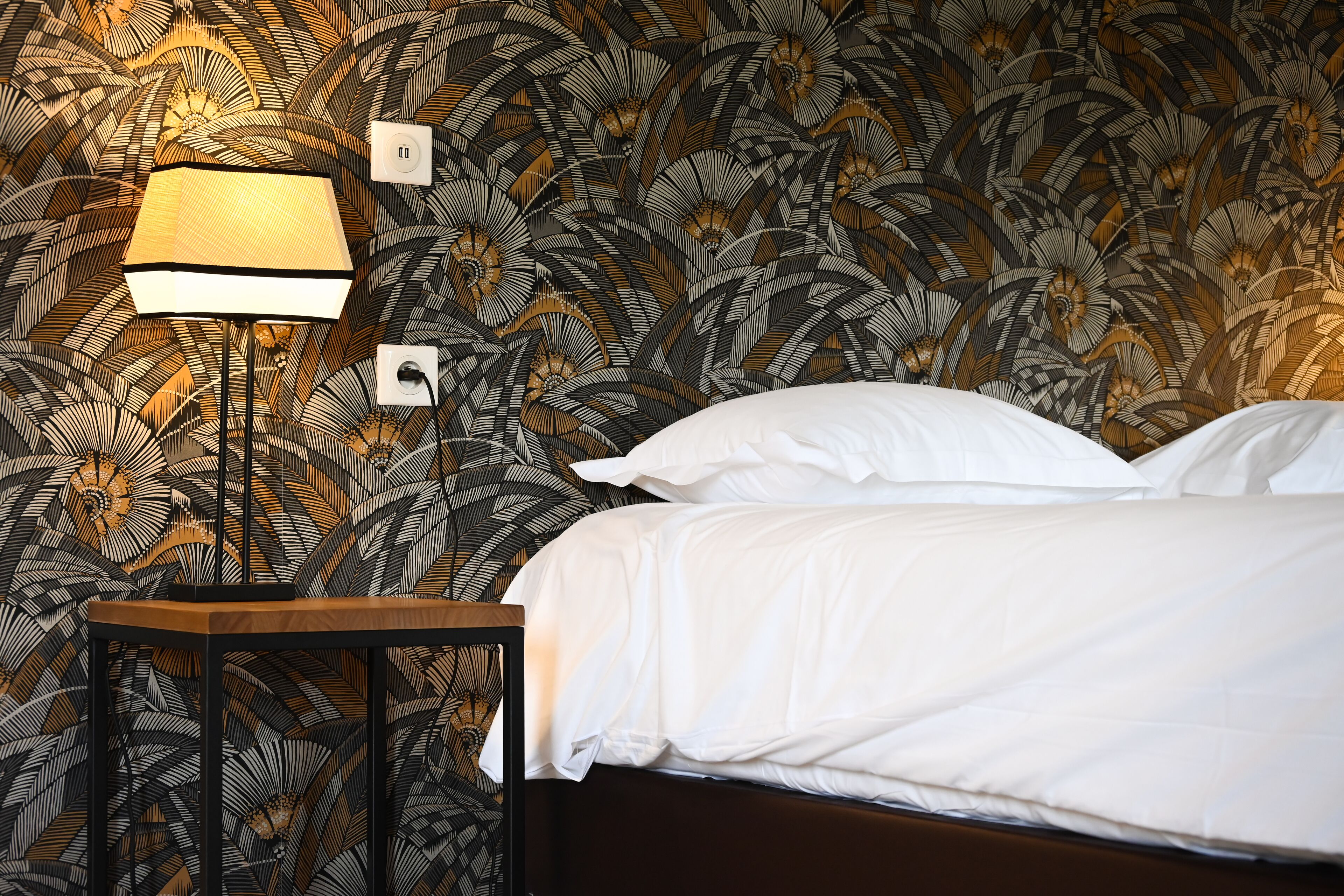A bedroom with a bed, white bedding, a nightstand with a lamp, and a patterned wallpaper.