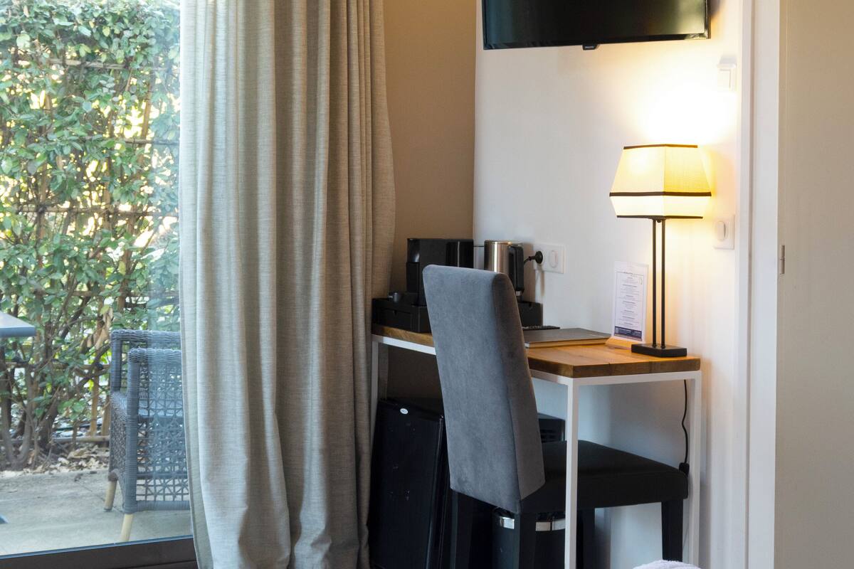 A hotel room with a bed, a desk with a chair, a TV mounted on the wall, and a window with curtains.