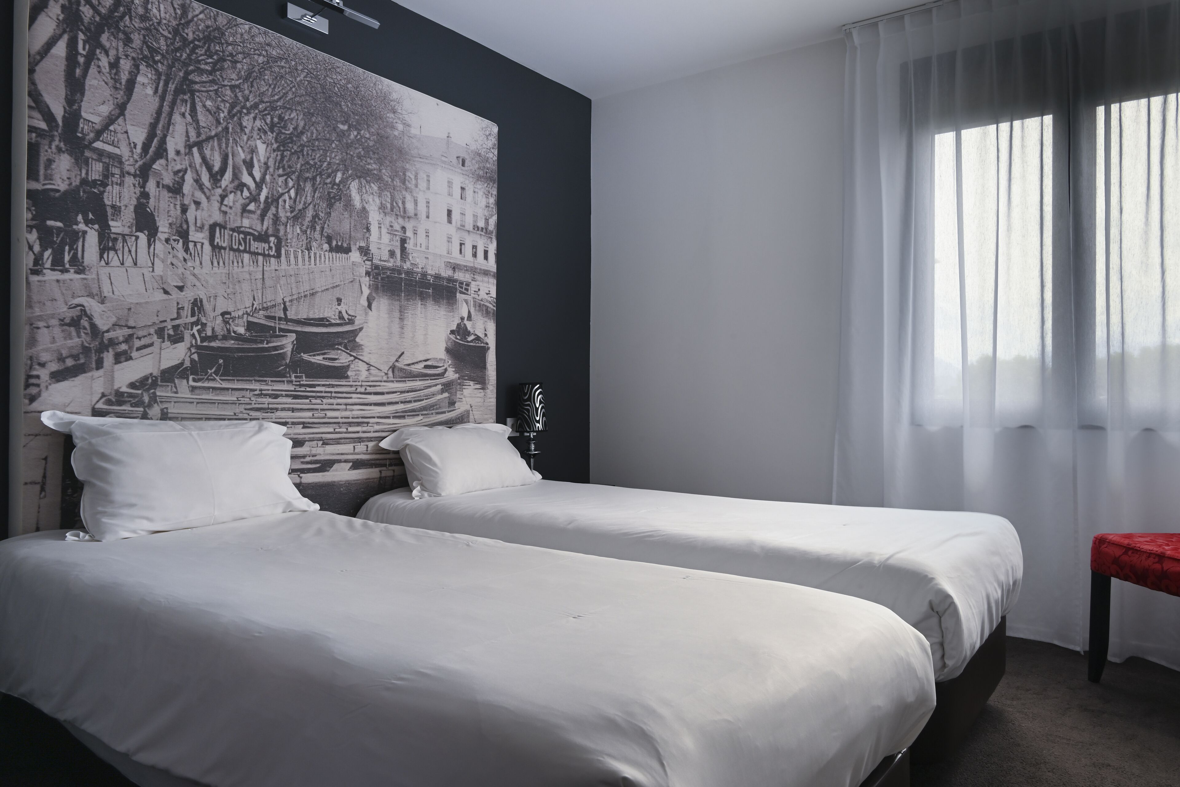 Una habitación de hotel con dos camas, un mural grande en blanco y negro en la pared, una ventana con cortinas translúcidas y un pequeño banco rojo.