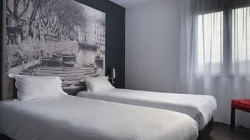 A hotel room with two beds, a large black and white mural on the wall, a window with sheer curtains, and a small red bench.