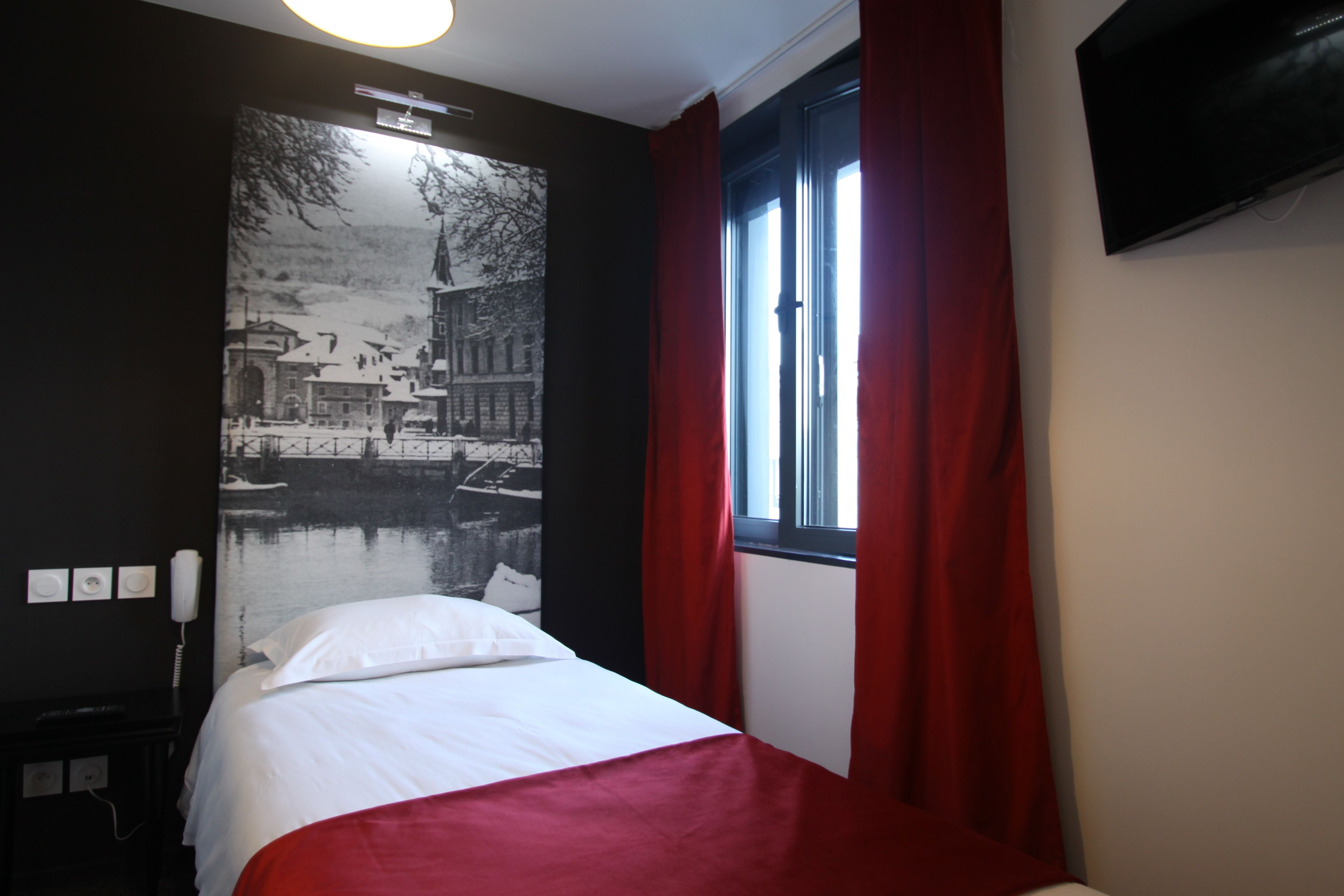 A hotel room with a bed, a large black and white photo on the wall, a window with red curtains, and a flat-screen TV mounted on the wall.