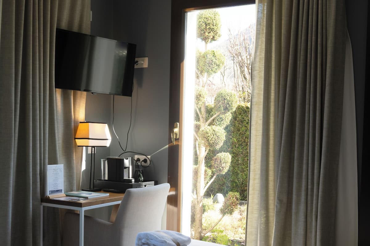 A hotel room with a bed, a desk with a TV, a lamp, and a view of trees.