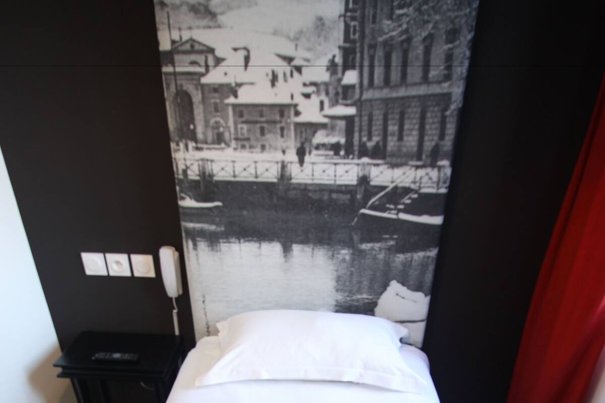 A bed with a red and white bedspread, a black nightstand with a lamp, and a black and white cityscape mural on the wall.