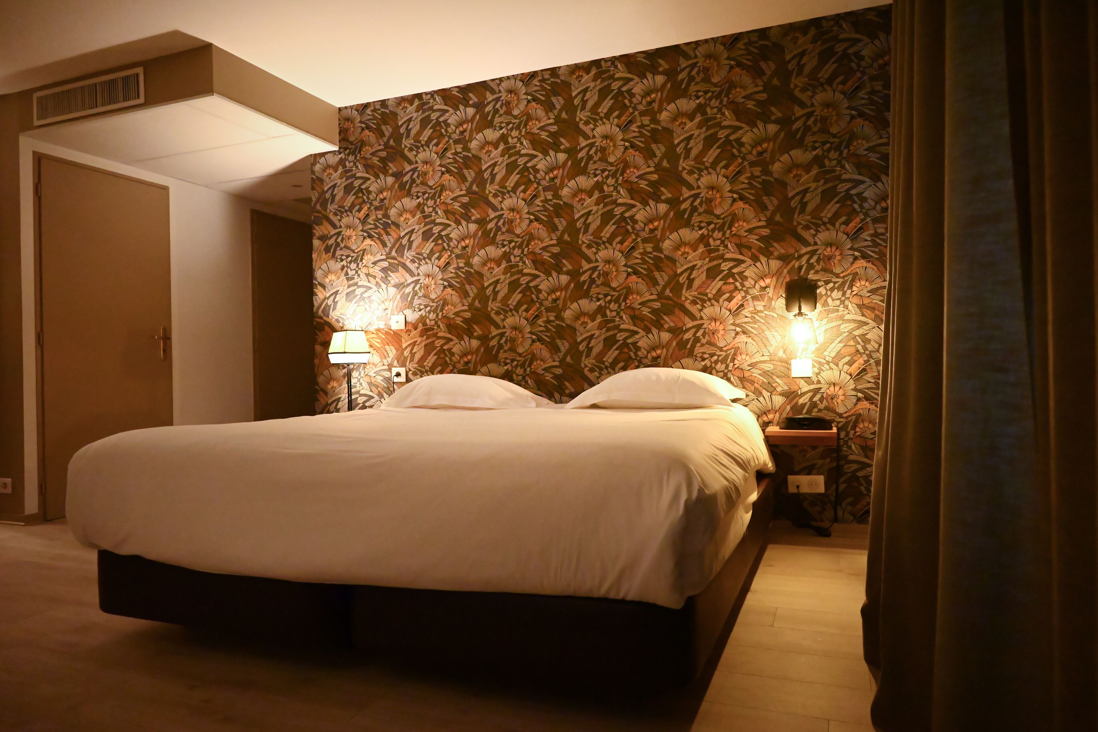 Una cama bien tendida en una habitación de hotel con papel tapiz floral, dos lámparas de noche y una puerta a otra estancia.
