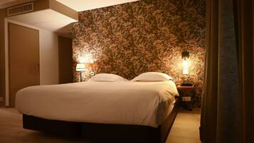Una cama bien tendida en una habitación de hotel con papel tapiz floral, dos lámparas de noche y una puerta a otra estancia.