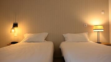 Una habitación de hotel con dos camas individuales, cada una con una colcha y almohadas blancas. Hay dos mesitas de noche, una al lado de cada cama, con lámparas encima. Las paredes tienen un diseño con textura.
