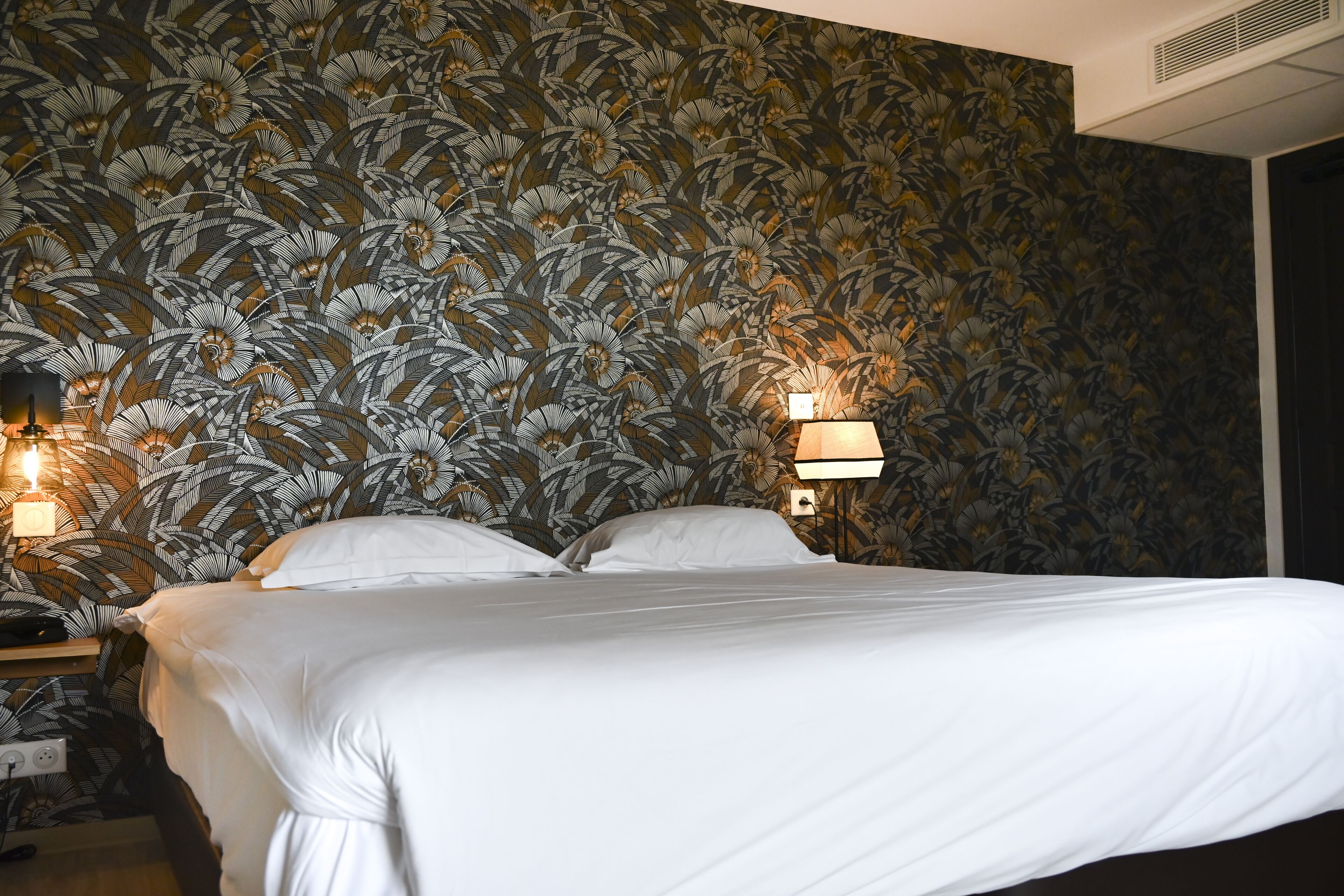 Una cama bien hecha con sábanas blancas en una habitación con papel tapiz floral y dos lámparas de pared.