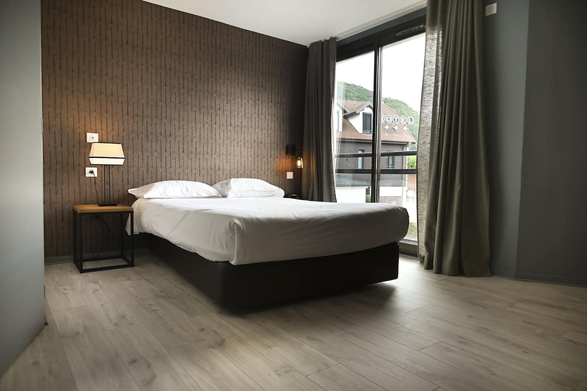 A modern hotel room with a large bed, a nightstand with a lamp, and a view of a building through a large window.