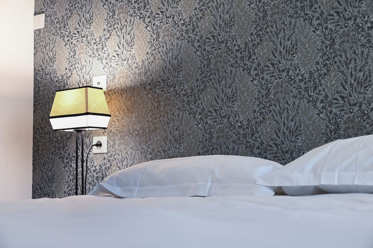 A hotel room with a patterned wallpaper, a bedside lamp, and a neatly made bed with white linens.