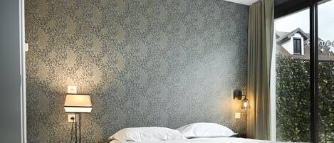 Un dormitorio con papel tapiz estampado, una cama con ropa de cama blanca, una mesita de noche con una lámpara y una ventana con cortinas.