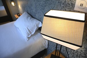 Una cama con ropa blanca y una lámpara con un diseño de rejilla sobre un soporte.
