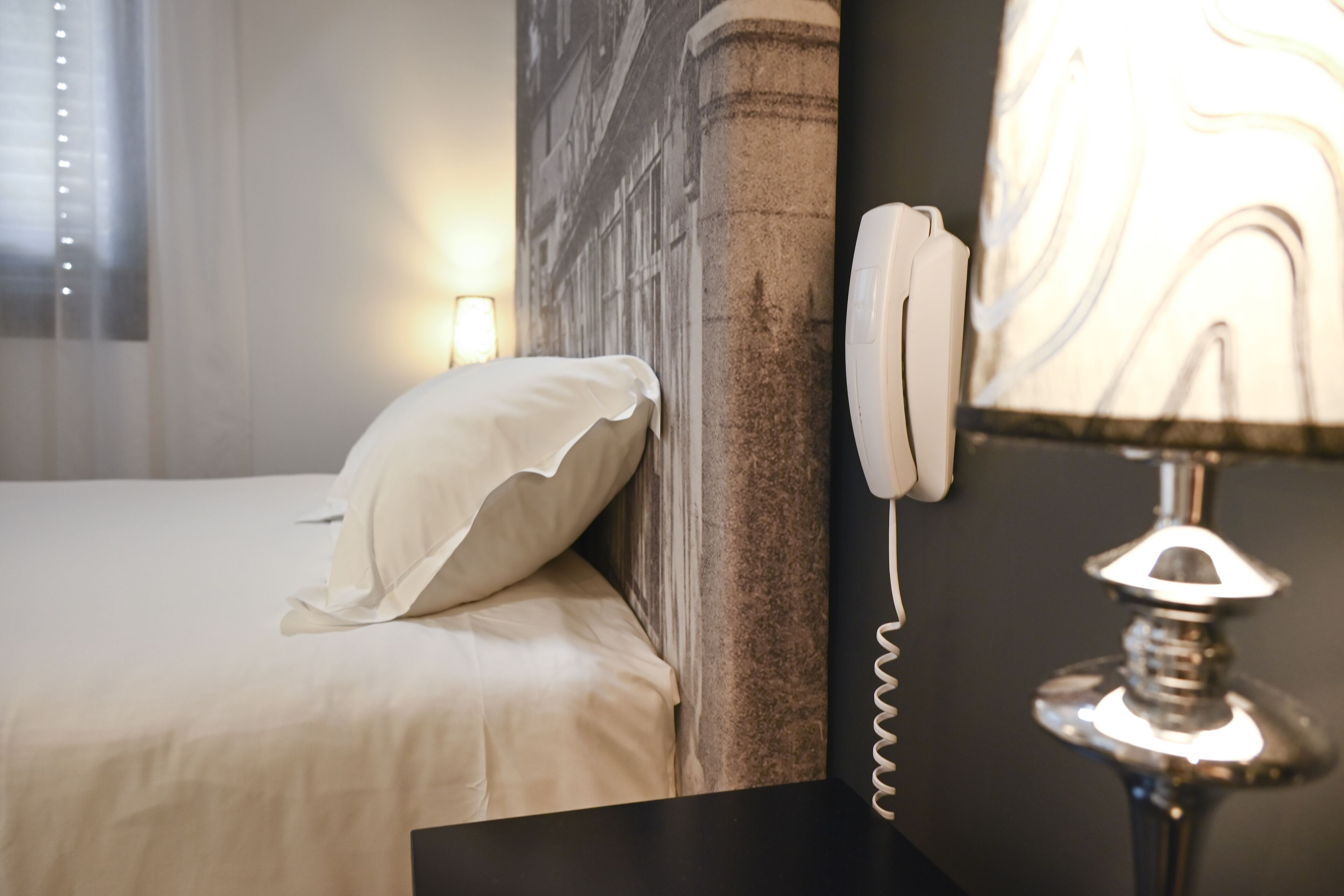 A neatly made bed with a pillow, a wall-mounted telephone, and a lamp with a marble-like shade.