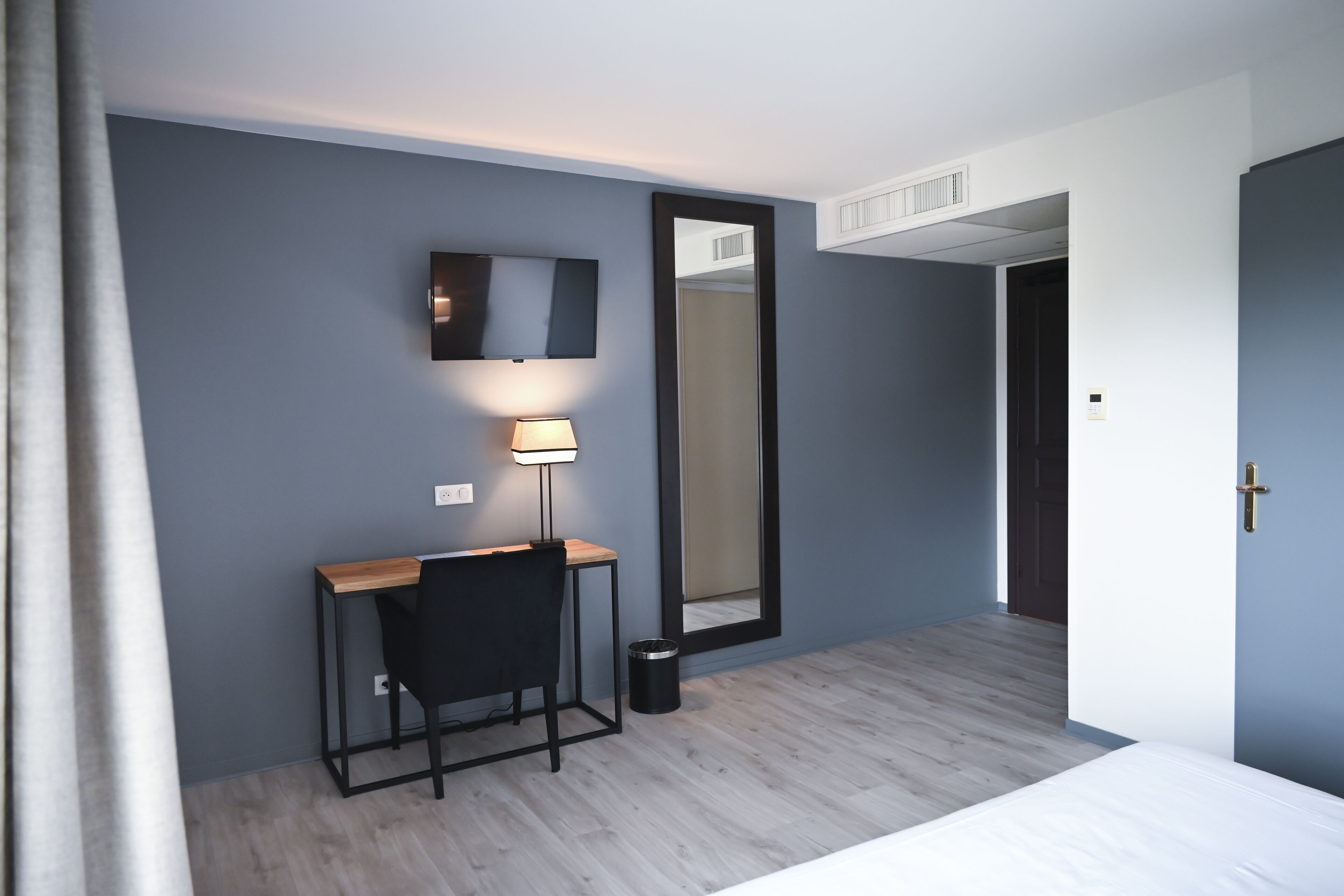 Una habitación de hotel con escritorio, silla, lámpara, espejo grande y una cama.