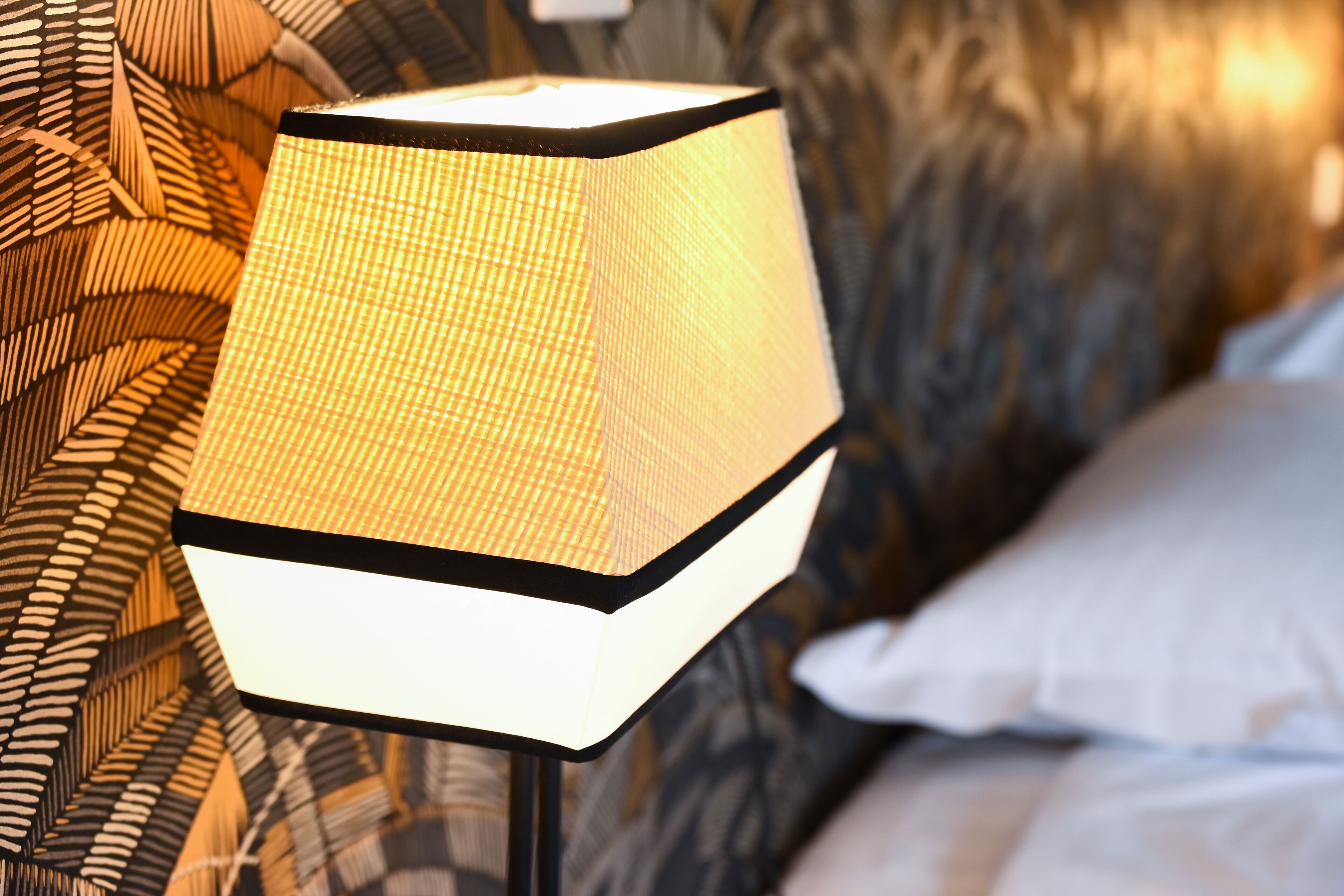 A lamp with a textured shade, a patterned wall, and a bed with a white pillow.