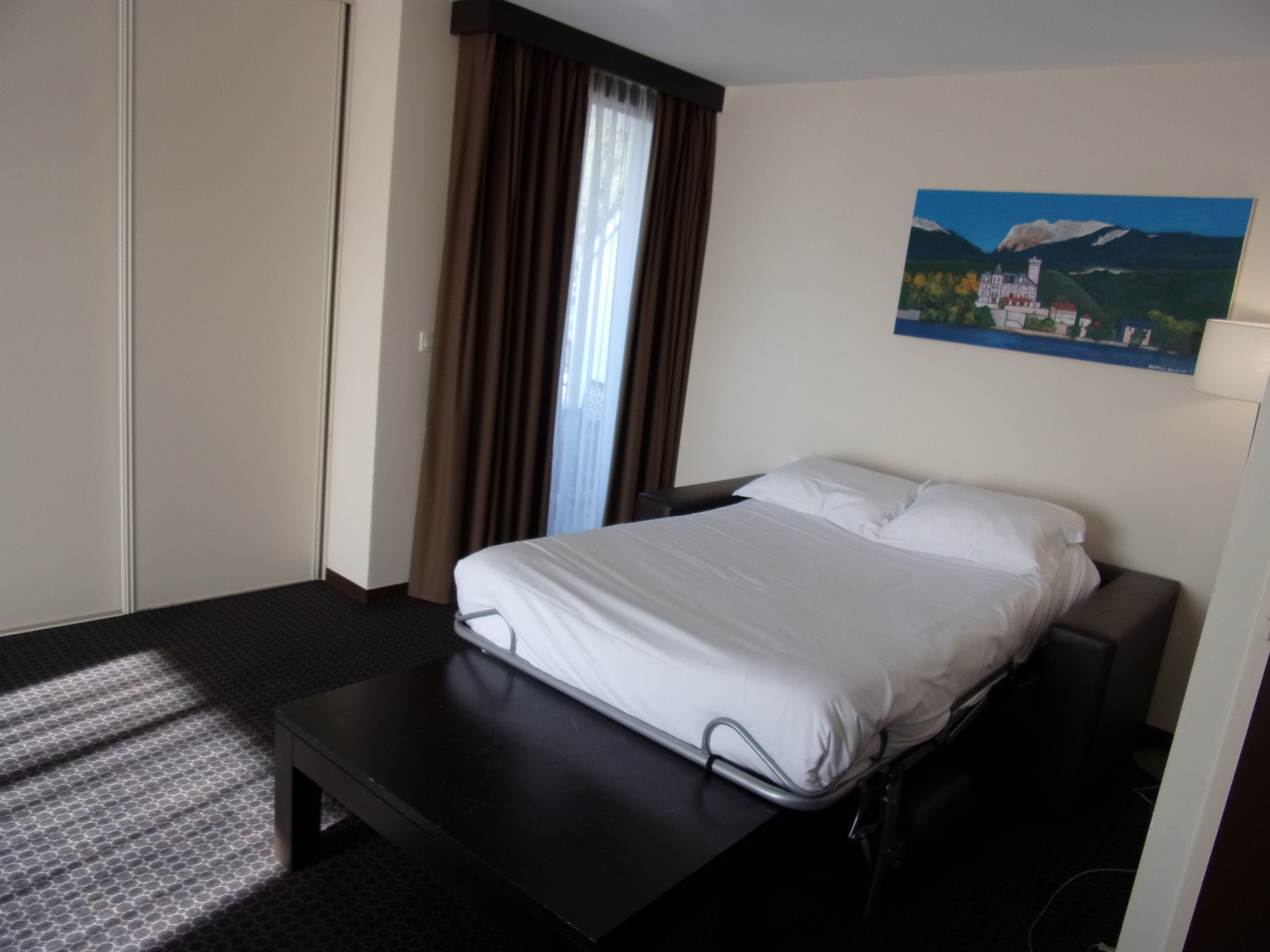 A hotel room with a bed, a desk, a painting on the wall, and a window with curtains.