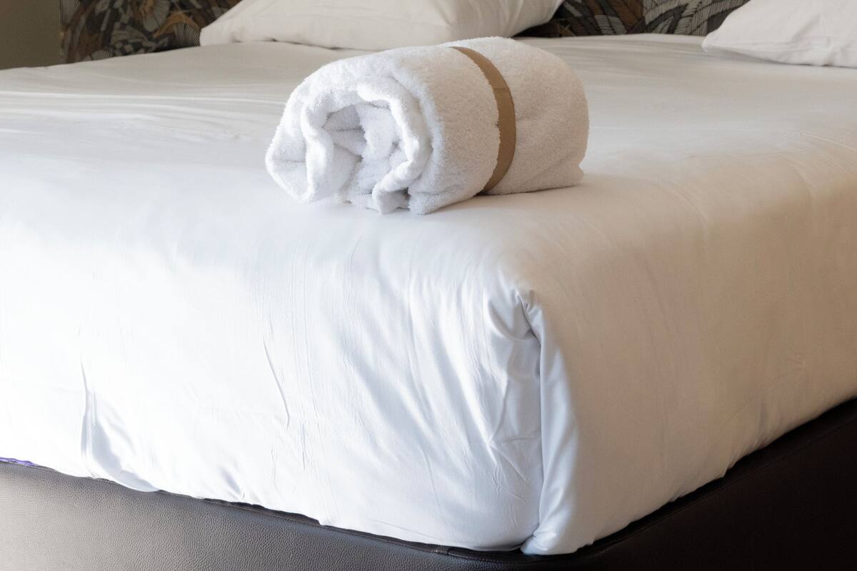 A neatly made bed with white linens and a rolled towel, a floral-patterned wall behind, and a black wall-mounted light fixture.