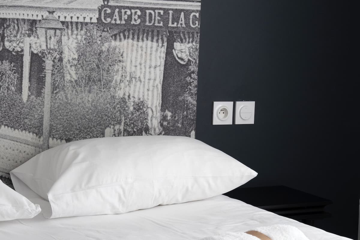 A neatly made bed with white linens and a rolled towel, a dark wall with a tapestry, and a sign reading 'HOTEL DE LA GA'.