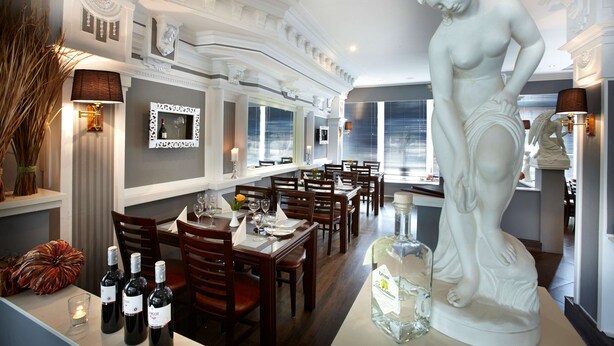 A dining area with wooden tables and chairs, a large white statue, and wine bottles. - Hotel Plöner See by Tulip Inn (Ploen)